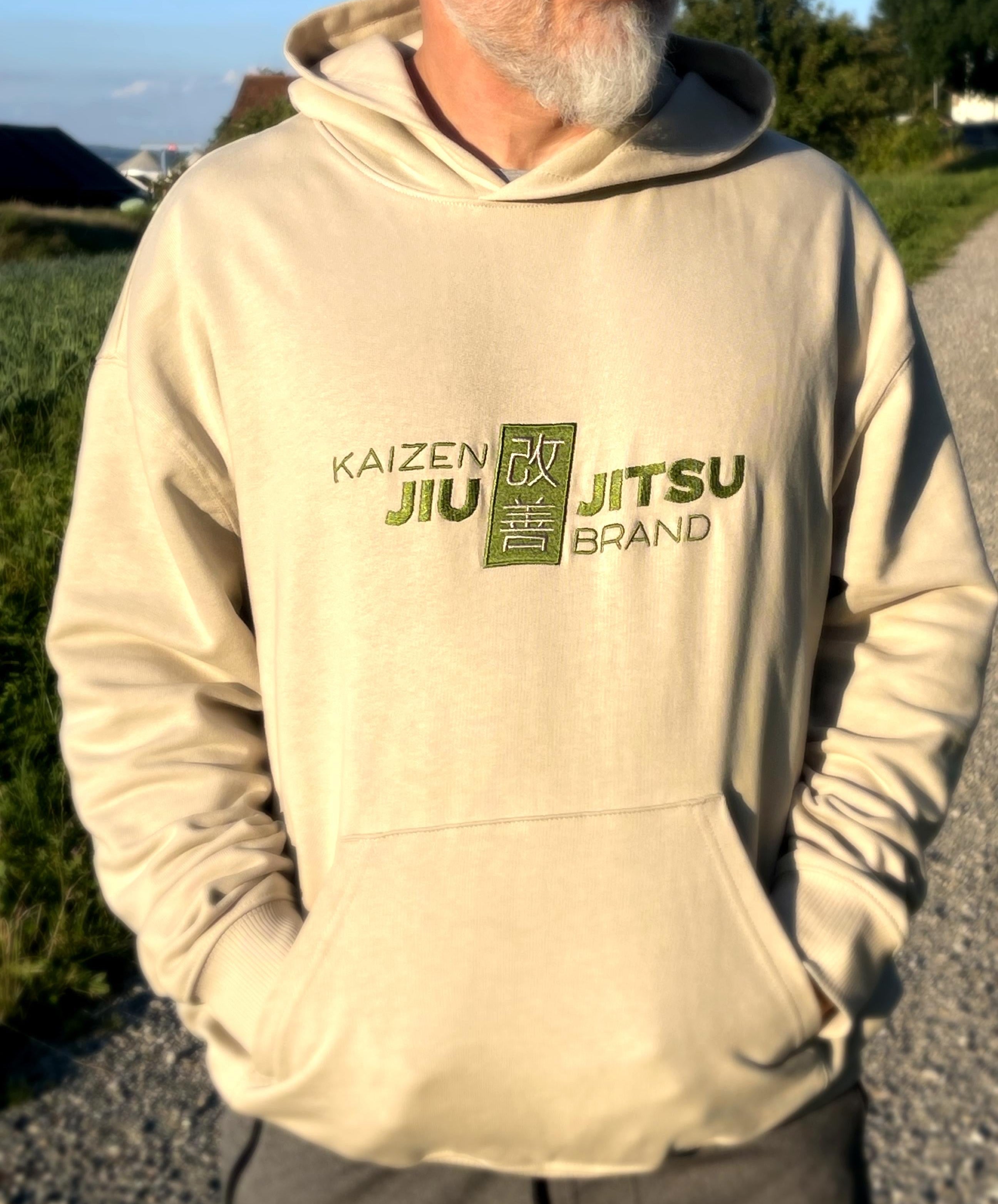 Kaizen Desert Dust Hoodie in Beige – Nahaufnahme des mossgrünen 改善 Jiu-Jitsu Brustlogos und der Kängurutasche