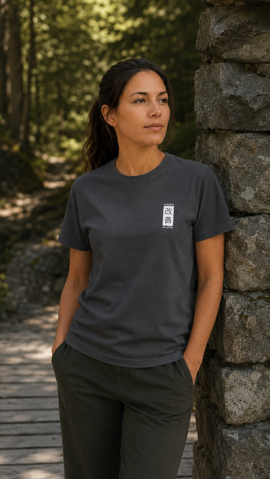 Frau im grauen Kaizen T-Shirt an einer Steinmauer im Wald – weisses Brustlogo, Frontansicht